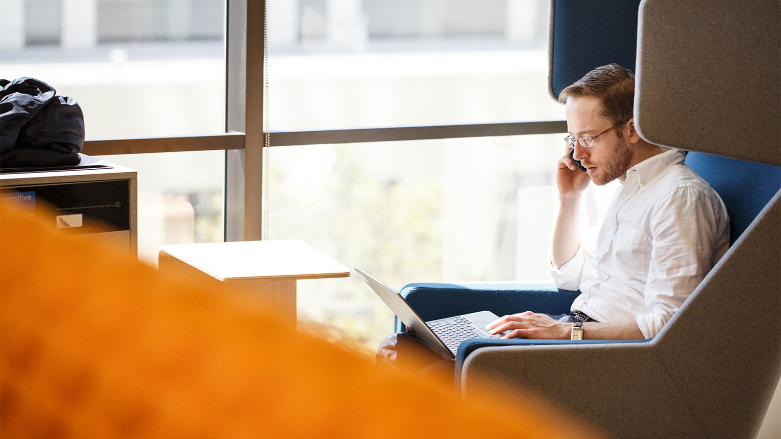 Man sitting on a chair talking on his cell phone while looking at his laptop Man sitting on a chair talking on his cell phone while looking at his laptop